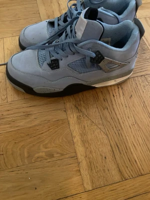 Jordan 4 University Blue sneakers - Snygga Jordan 4 sneakers i färgen University Blue med ovandel i mocka och detaljer i svart och grått. Skorna har meshpaneler på sidorna, klassisk snörning och den ikoniska Jumpman-loggan på hälen. Perfekta för dig som vill sticka ut med stilrena och sportiga sneakers.