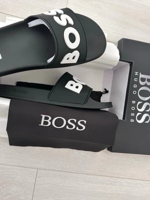 Svarta BOSS sandaler med logga - Svarta sandaler från Hugo Boss med stor vit BOSS-logga över foten. Tillverkade i syntetmaterial med räfflad sula för extra grepp. Kommer med originalförpackning och dustbag. Perfekta för en chill och stilren look. 46 men passar mer 45
