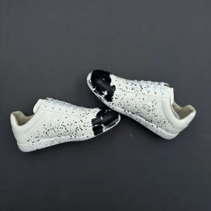 Vita sneakers från Maison Margiela med svart färgstänk - Unika vita sneakers från Maison Margiela i läder med svart färgstänk. Klassisk låg modell med vita skosnören och diskret logotyp på sulan. Perfekt för dig som gillar statement-plagg och streetstyle.