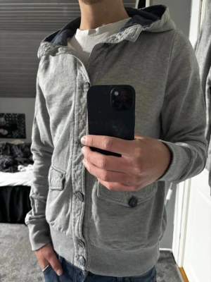 Grå hoodie - Snygg grå hoodie med knäppning framtill och två stora fickor med lock. Huvan har mörkare innerfoder och tröjan har ribbade muddar vid ärmslut och nederkant. Perfekt för en avslappnad och stilren look.
