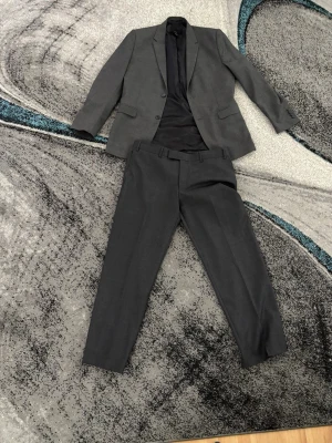 Mörkgrå kostym set - Snygg mörkgrå kostym från H&M med klassisk tvåknäppt kavaj och matchande byxor. Kavajen har smal passform, två fickor med lock och en bröstficka. Perfekt för dig som vill ha en stilren och enkel look.                                                               Helt nya använda en gång                                                Byxorna:97/76.             Kavaj:54