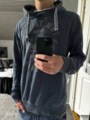 Tom Tailor vintage - Snygg blå hoodie från Tom Tailor med tryck framtill och vita dragsnören i huvan. Hoodien har ribbade muddar vid ärmslut och nederkant samt en avslappnad passform. Perfekt för en casual look.
