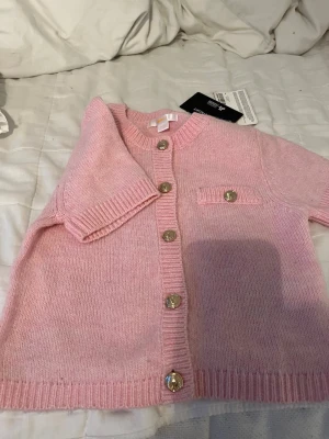 Ljusrosa/ kort t shirt  kofta från Gina Tricot - Supersöt ljusrosa kofta från Gina Tricot med guldfärgade knappar och rund halsringning. Koftan har ribbade muddar, en liten dekorficka och är stickad i mjukt material. Perfekt för att addera en gullig touch till din outfit.