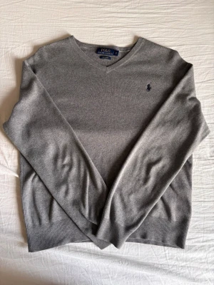 Grå V-ringad tröja från Polo Ralph Lauren - Snygg grå V-ringad tröja från Polo Ralph Lauren med den klassiska broderade loggan på bröstet. Tröjan har ribbade muddar vid ärmslut och nederkant, och är tillverkad i ett mjukt och tunt material som passar perfekt till lager-på-lager.