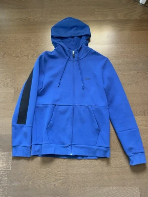 Schysst blå zip hoodie från Boss i gott skick - Snygg blå hoodie från Boss med dragkedja framtill och huva med dragsko. Ena ärmen har en svart rand som ger en sportig touch. Hoodien har två fickor fram och diskret Boss-logga på bröstet. Perfekt för en avslappnad och trendig stil. Tröjan kosta från början 2000 och är nästan helt oanvänd, fick den i present men är inte min still riktigt..