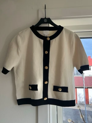 Crèmevit kofta med guldknappar - Kortärmad vit kofta med svarta kanter och dekorativa guldknappar framtill. Två små fickor framtill med svarta lock och guldknapp. Rund halsringning och rak passform.