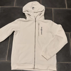 Vit hoodie från Sail Racing - Snygg vit hoodie från Sail Racing i storlek 170, med dragkedja framtill och logga på bröstet. Hoodien har justerbar huva med snören och en liten logga även på huvan. Den är ny utan prislapp så aldrig använt!