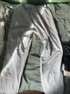 Ljusgrå mjukisbyxor från Under Armour - Säljer ett par ljusgrå mjukisbyxor från Under Armour med resår i midjan och logga på vänster lår. Byxorna har en avslappnad passform och är tillverkade i ett lätt och bekvämt material, perfekta för chill dagar eller träning.
