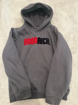 Grå HOODRICH hoodie med brodyr - Snygg grå hoodie från HOODRICH med broderad logga i rött och svart på bröstet. Klassisk modell med huva och ribbade muddar vid ärmar och nederkant. Perfekt för en avslappnad streetwear-look.