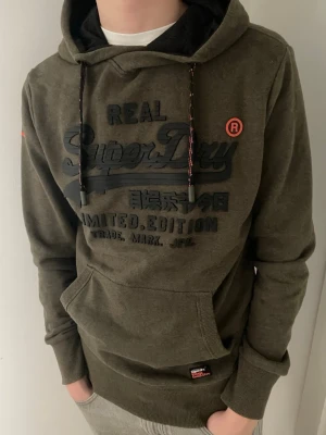 Olivgrön hoodie från Superdry - Snygg olivgrön hoodie från Superdry med stor logga och text på bröstet. Hoodien har huva med röda och vita snören samt en klassisk känguruficka framtill. Tillverkad i mjuk bomullsblandning och har ribbade muddar vid ärmslut och nederkant. Det står storlek M men den är ganska liten i  storlek så passar bättre som storlek S. Modellen är 180 och väger 65