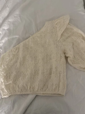 Vit spetsig offshoulder topp - Säljer en superfin vit offshoulder topp med spetsdetaljer och broderat mönster. Toppen har en puffig ärm på ena sidan och är croppad i modellen. Perfekt för dig som gillar romantisk och trendig stil.