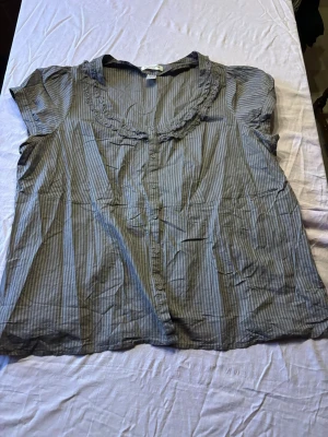 Gråblå randig blus med volang - Snygg gråblå blus med tunna vita ränder och volangdetalj runt den runda halsringningen. Blusen har knäppning framtill och korta ärmar. Tillverkad i ett lätt och luftigt tyg, perfekt för en avslappnad stil.