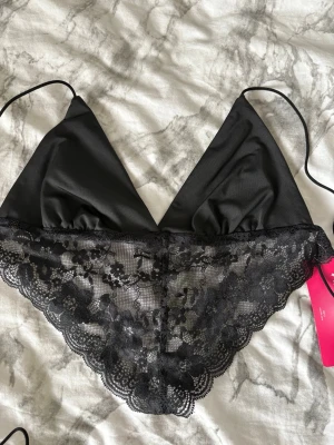 Svart spetstopp med tunna band - En svart spetstopp med tunna axelband och triangelformade kupor. Nedre delen är dekorerad med vacker blommig spets som ger en romantisk känsla. Materialet är mjukt och lätt, perfekt för en bekväm känsla. Lappen sitter kvar.