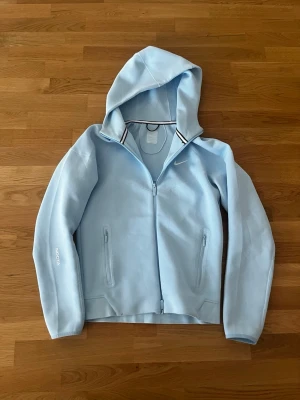 Ljusblå hoodie från Nike NOCTA - Snygg ljusblå hoodie från Nike NOCTA med dragkedja framtill och två fickor med dragkedja. Hoodien har en klassisk huva och diskret Nike-logga på bröstet samt NOCTA-tryck på ärmen. Tillverkad i mjukt material som känns skönt mot huden.