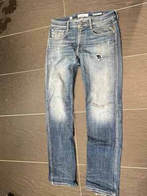 Blå slitna jeans från Replay  - Säljer ett par blå jeans från Replay med slitningar och hål på framsidan för en cool, avslappnad look. Klassisk femficksmodell med raka ben och ljusa tvättade partier. Perfekta för dig som gillar streetstyle och vill ha ett par jeans med attityd. W32 L34. Modell: anbass 05 aged