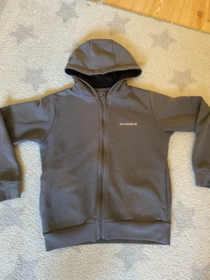 Grå zip hoodie från McKenzie - Snygg grå hoodie från McKenzie med dragkedja framtill och huva. Tröjan har ribbade muddar vid ärmslut och nederkant samt diskret logga på bröstet. Perfekt för en avslappnad och stilren look.