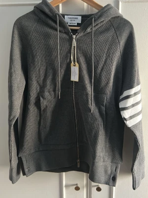 Grå zip hoodie från Thom Browne - Jag säljer en riktigt snygg tröja från Thom Browne som jag säljer då den är lite för liten för mig tyvärr. Helt nytt skick och köpt på TB’s hemsida och originalpåsen finns kvar.