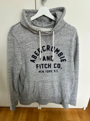 Grå hoodie från Abercrombie & Fitch - Mysig gråmelerad hoodie från Abercrombie & Fitch med stor logga i mörkblått framtill. Klassisk huva med vita dragsnören och känguruficka. Tillverkad i mjukt material som passar perfekt för en avslappnad stil.