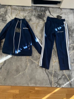 Palm Angels tracksuit - Snyggt blått tracksuit-set från Palm Angels med vit logga på både jacka och byxor. Setet har vita ränder längs ärmar och ben, dragkedja på jackan och elastisk midja på byxorna. Perfekt för en sportig och trendig look. Pris kan ev disskuteras, enbart seriösa köpare. Kan vid efterfrågan även mötas i Stockholm. Allt är i nyskick