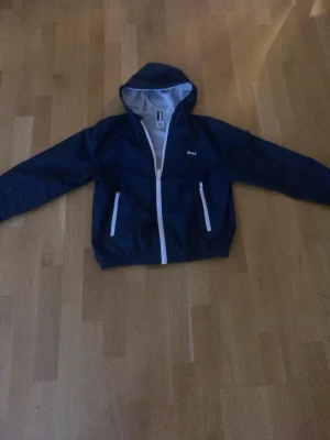 Marinblå vindjacka från BOSS - Snygg marinblå vindjacka från BOSS med vit dragkedja och vita detaljer. Jackan har huva, två fickor med dragkedja och meshfoder på insidan. Perfekt för blåsiga dagar och enkel att matcha till olika outfits.