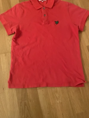 Röd pikétröja från Lyle & Scott - Klassisk röd pikétröja från Lyle & Scott med broderad grön logga på bröstet. Tröjan har korta ärmar, krage och knappar framtill. Perfekt till en avslappnad och stilren look.
