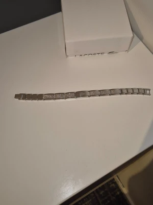 Silverfärgat armband från Lacoste - Snyggt armband i silverfärgad metall från Lacoste. Länkarna har en mix av slät och texturerad yta, och mittdelen pryds av den klassiska Lacoste-loggan. Perfekt accessoar för dig som gillar stilrena detaljer och sportig känsla. Nypris 1k nått mitt pris 549.