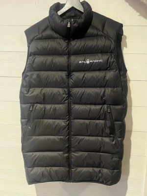 Svart dunväst från Sail Racing - Snygg svart dunväst från Sail Racing med hög krage och dragkedja framtill. Västen har två fickor med dragkedja och logga på bröstet samt en diskret logga på ryggen. Perfekt för lager-på-lager och håller dig varm under kyliga dagar.