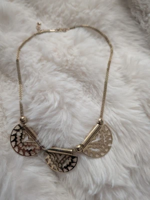 Guldigt halsband med halvcirkelhängen - Unikt halsband i guldfärgad metall med tre halvcirkelformade hängen med utskurna mönster. Kedjan är tunn och elegant, och hänget har detaljerade geometriska former som ger ett coolt och trendigt intryck. Perfekt för dig som gillar statement-smycken.