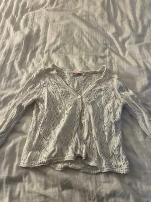 Vit spetskofta med knappar - Söt vit kofta i spetsmönster med v-ringning och knäppning framtill. Perfekt att ha över badkläder på stranden eller i vardagen på sommarn.