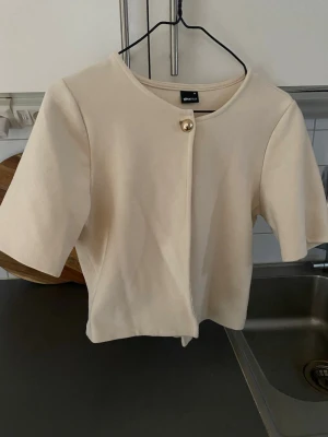 Krämvit kortärmad kofta Gina Tricot - Stilren krämvit kofta från Gina Tricot med korta ärmar och rund halsringning. Koftan har en dekorativ guldig knapp framtill och är gjord i ett mjukt, stretchigt material. Perfekt att slänga över en topp för en clean look.