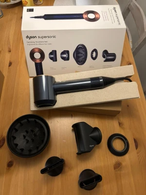 Dyson Supersonic hårfön med tillbehör - Säljer en Dyson Supersonic hårfön i mörkgrå metallic med kopparfärgad ring. Medföljer flera munstycken och diffuser, allt i originalförpackning. Modern design och tillverkad i plast och metall. Perfekt för dig som vill ha snabb och skonsam hårtorkning.