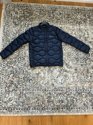 Mörkblå dunjacka från PeakPerformance - Snygg mörkblå dunjacka från PeakPerformance med quiltat mönster och snygg krage. Jackan har dragkedja framtill och två sidofickor. Perfekt för kalla dagar och enkel att matcha med olika outfits. Jackan har endast några få blå märken/ärr som du ser på den sista bilden. Annars är den i väldigt fin skick och jag har endast använt den ett fåtal gånger.