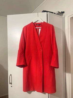 Röd kappa från H&M med knappar - Stilren röd kappa från H&M med rak passform och två knappar framtill. Kappan har diskreta fickor på sidorna och en enkel, kraglös design som ger en modern look. Perfekt för att addera färg till din outfit.
