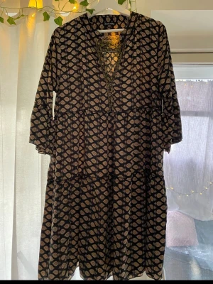 Svart mönstrad klänning Black Colour - Svart klänning från Black Colour med beige grafiskt mönster, trekvartsärm. Klänningen har en lös passform och faller strax ovanför knäna. Perfekt för dig som gillar boho-vibbar och snygga detaljer.