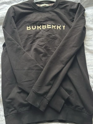 Svart sweatshirt från Burberry - Säljer en svart sweatshirt från Burberry med broderad logga och text på bröstet. Klassisk rund halsringning och ribbade muddar vid ärmslut och nederkant. Insidan av kragen har det ikoniska Burberry-mönstret. Perfekt för en stilren och lyxig look. 