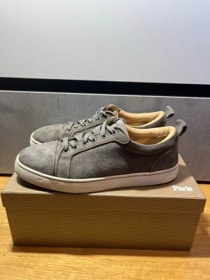 Gråa sneakers från Christian Louboutin - Stilrena gråa sneakers i mocka från Christian Louboutin med klassisk röd sula och beige innersida. Lågt skaft och vita sulor ger en clean look. Perfekta för dig som vill ha något exklusivt och tidlöst i garderoben. Ställ gärna frågor eller prisförslag! Skickar inom MAX 2 dagar [Oftast 1 dag]!