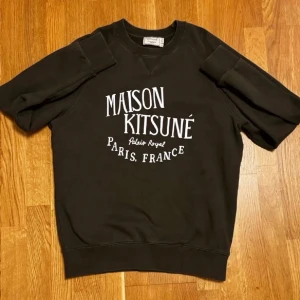 Maision Kitsuné Tröja - Olivgrön sweatshirt från Maison Kitsuné i storlek L, men sitter mer som M. Detta är en populär modell i en mer sällsynt färg. Tröjan har korta ärmar och tryck med texten “Maison Kitsuné Paris, France” framtill. Tillverkad i Portugal av 100 % bomull.