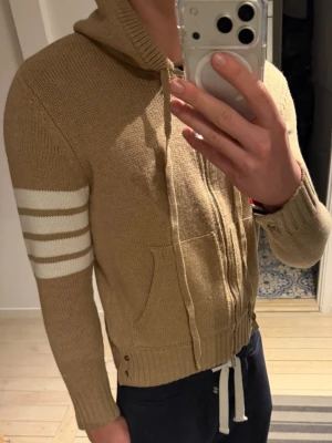 Beige stickad hoodie med dragkedja - Säljer en beige stickad hoodie med dragkedja och vita ränder på ena ärmen. Hoodien har fickor framtill, ribbade muddar och dragsko i midjan. Perfekt för dig som gillar en avslappnad och sportig stil.