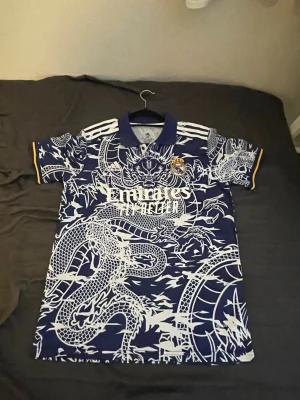 Real Madrid drakmönstrad matchtröja - Säljer en blå Real Madrid matchtröja från Adidas med unikt vitt drakmönster över hela tröjan. Tröjan har korta ärmar, gul detalj på ärmslut och klubbmärke på bröstet. Kragen är blå och modellen är rak. Perfekt för dig som vill sticka ut på planen.