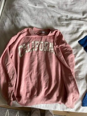 Rosa sweatshirt CALIFORNIA - Säljer en rosa sweatshirt med trycket 'CALIFORNIA' i stora vita bokstäver över bröstet. Tröjan har rund halsringning och ribbade muddar vid ärmslut och nederkant. Perfekt för en avslappnad och cool stil.