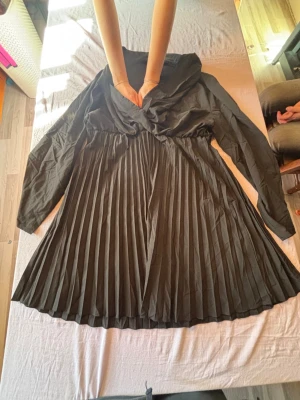 Svart plisserad klänning med v-ringning - Snygg svart klänning med v-ringad omlottdesign och långa ärmar. Kjoldelen är plisserad vilket ger ett elegant fall. Perfekt för dig som gillar stilrena och klassiska plagg med en twist.