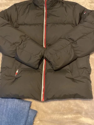 Svart pufferjacka från Tommy Hilfiger - Snygg svart pufferjacka från Tommy Hilfiger med dragkedja framtill och diskreta röda och vita detaljer på dragkedjan och fickorna. Jackan har hög krage och klassisk quiltad design. Perfekt för kalla dagar och riktigt stilren.