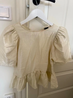 Beige puffärm blus med volang - Trendig beige blus med puffärmar och volang nedtill. Tillverkad i ett glansigt material som ger en lyxig känsla. Perfekt för dig som vill sticka ut med en feminin och modern look.