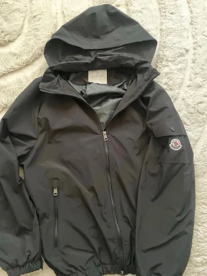 INTRESSEKOLL Svart vindjacka från Moncler - INTRESSEKOLL.       Snygg svart vindjacka från Moncler med huva och dragkedja framtill. Jackan har elastiska muddar vid ärmslut och nederkant, samt en ficka med knapp på ärmen där Monclers logga sitter. Perfekt för blåsiga dagar.