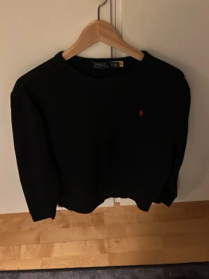 Svart sweatshirt från Polo Ralph Lauren - Klassisk svart sweatshirt från Polo Ralph Lauren med rund halsringning och den ikoniska röda logotypen broderad på bröstet. Tröjan har en avslappnad passform och är tillverkad i mjukt material som passar perfekt till jeans eller chinos.