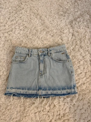 Ljusblå jeanskjol med fransig kant - Säljer en ljusblå somrig jeanskjol. Kjolen har bälteshällor, knapp och dragkedja framtill. Perfekt för dig som gillar en avslappnad och trendig stil. Den är köpt från Pull & Bear och har inga defekter. 