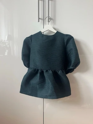 Mörkgrön kort klänning med puffärmar - Unik mörkgrön kortklänning med voluminösa puffärmar och markerad midja. Klänningen har rund halsringning och snygg knytning i ryggen som ger extra detalj. Materialet är strukturerat och ger en exklusiv känsla. Perfekt för dig som vill ha en statement-look.