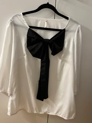 Vit blus med stor svart rosett - Snygg vit blus med en stor svart rosett framtill som ger en trendig look. Blusen har trekvartsärm och är gjord i ett lätt, mjukt tyg som känns skönt mot huden. Perfekt för dig som vill sticka ut med en unik detalj.