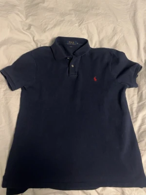 Mörkblå piké från Polo Ralph Lauren - Klassisk mörkblå pikétröja från Polo Ralph Lauren med röd broderad logga på bröstet. Skjortan har korta ärmar, krage och knappar framtill. Tillverkad i mjuk bomull som känns skön mot huden. Perfekt för en clean och stilren look.