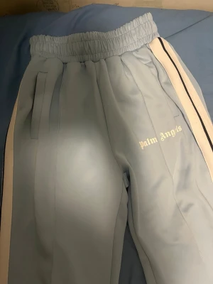 Palm Angels ljusblå mjukisbyxor - Säljer ett par ljusblå mjukisbyxor från Palm Angels med vita och svarta ränder längs sidorna. Byxorna har resår i midjan, raka ben och sidofickor. Palm Angels-loggan är tryckt i vitt på ena benet. Perfekta för en chill och trendig look.